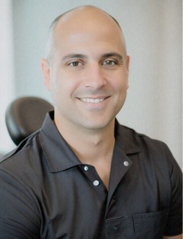 A person in a black shirt smiles while seated indoors. The background is neutral, with a visible chair. No landmarks or historical buildings present.