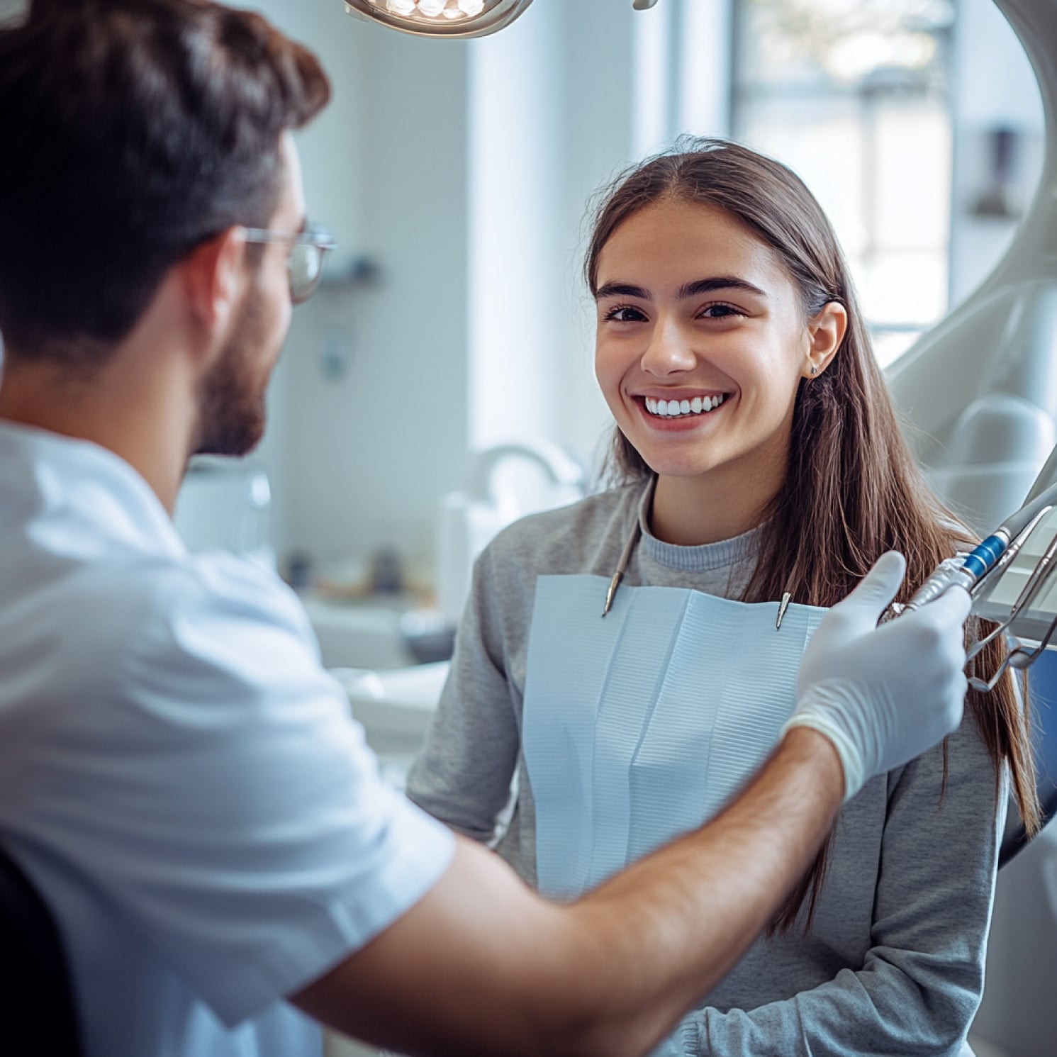 A person sits in a dental office, smiling, while another person holds dental equipment, under bright lights in a modern setting.