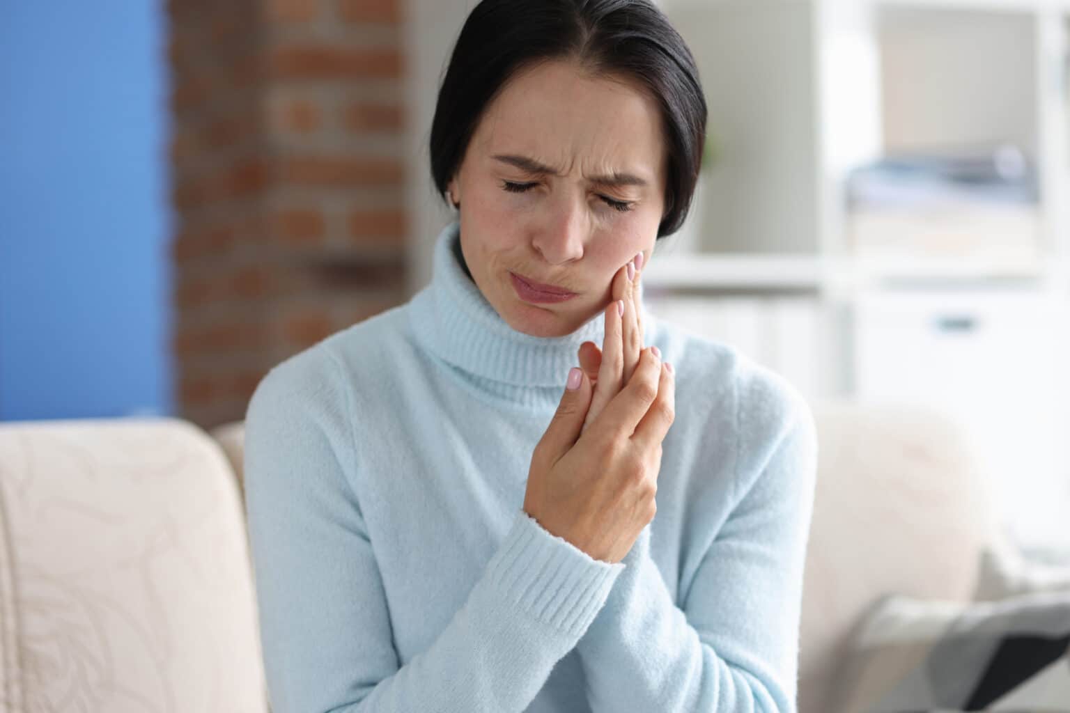 A person in a light blue sweater looks distressed, holding their cheek in discomfort, likely due to a toothache.