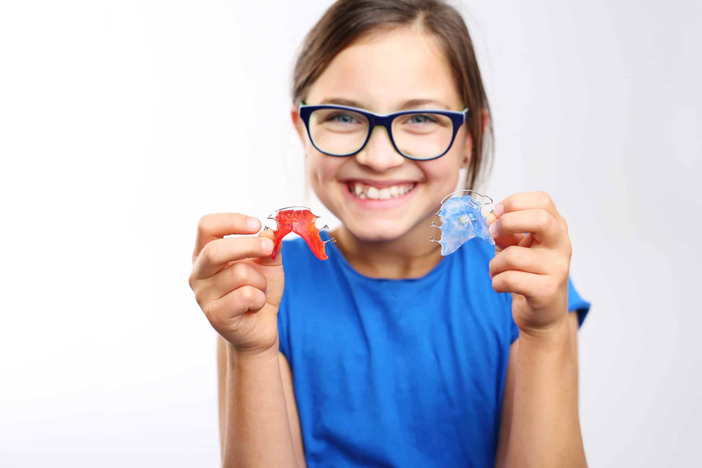 A smiling child with glasses holds a red retainer in one hand and a blue retainer in the other, wearing a blue shirt.