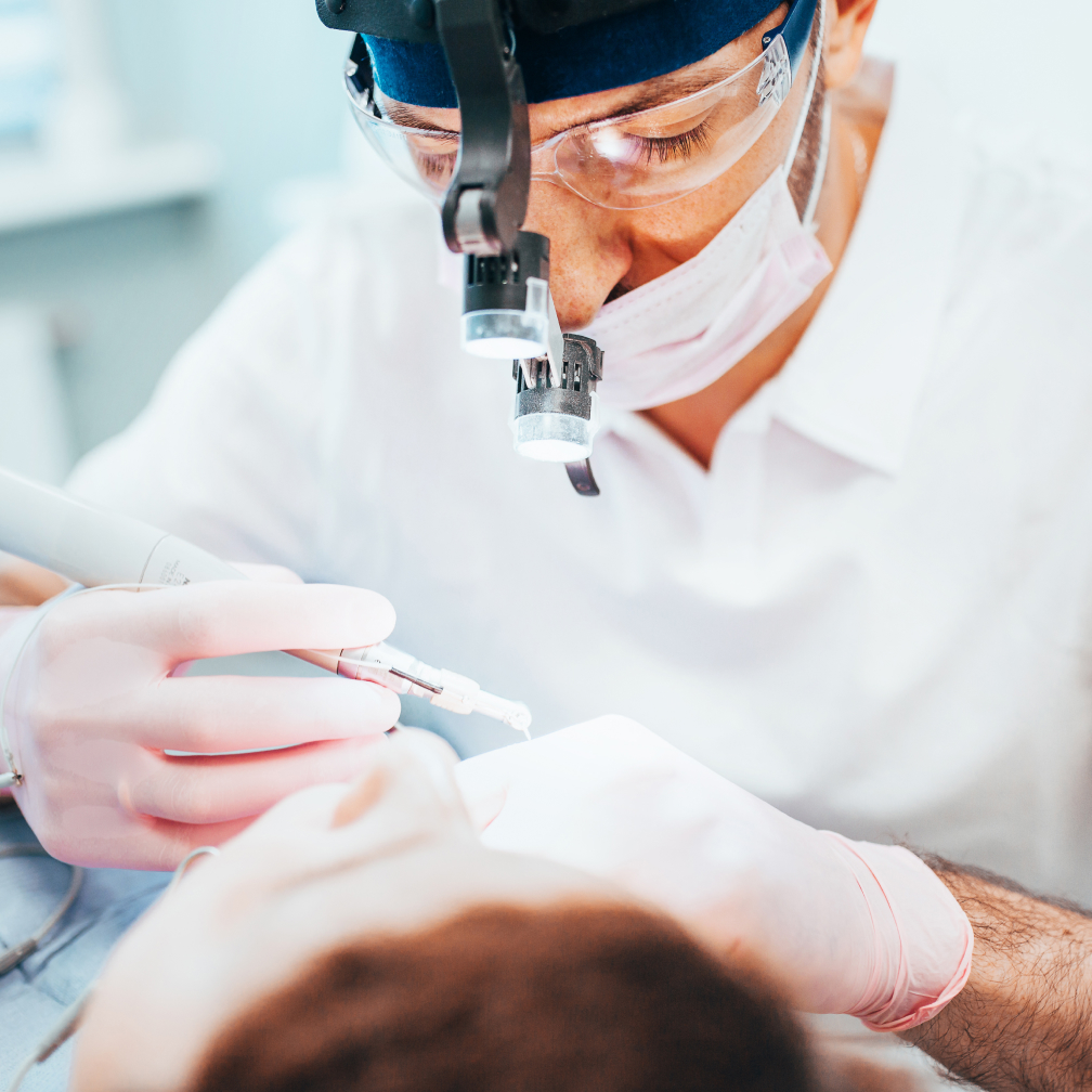 A person wearing a headlamp and protective gear performs dental work on a patient, focusing with precision instruments in a clinical setting.