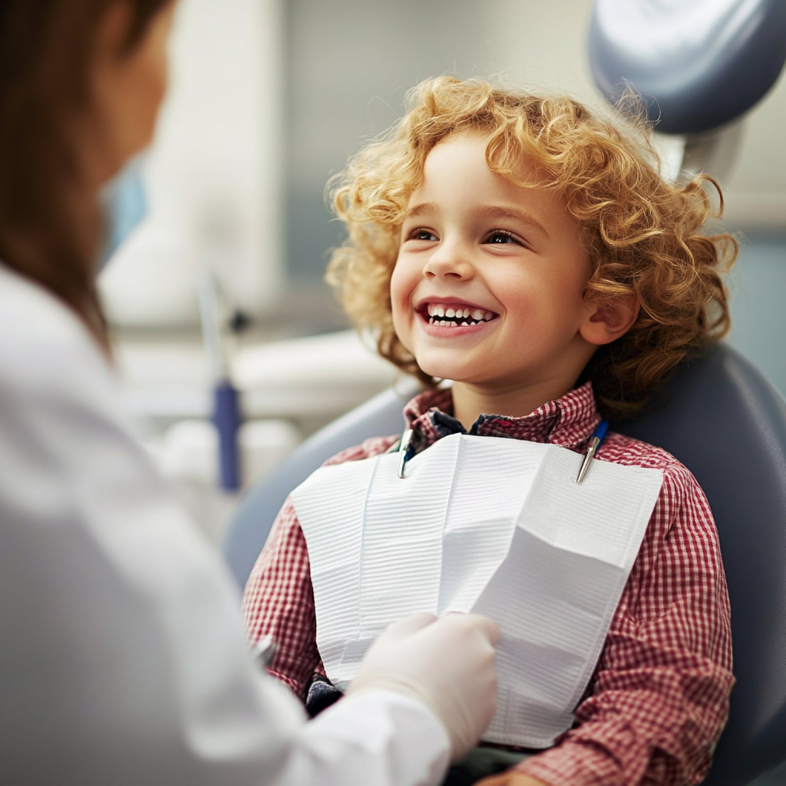 A child with curly hair smiles at a person in a dental office, wearing a checkered shirt and dental bib, feeling relaxed.